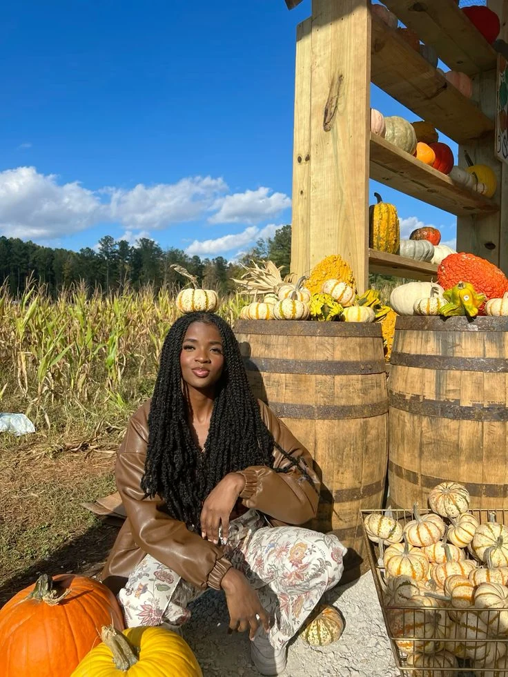 Woman in brown leather bomber jacket and floral pants crouching by pumpkin barrels at a fall farm