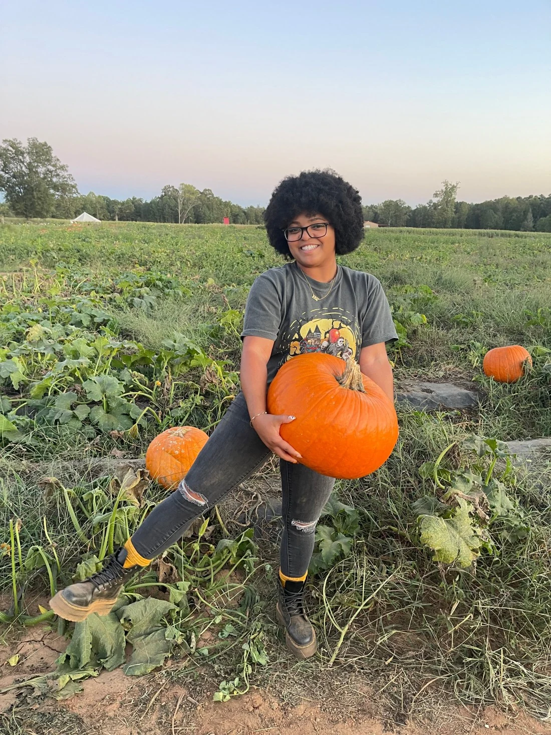 Woman in graphic tee, ripped jeans and combat boots holding a pumpkin in a pumpkin patch