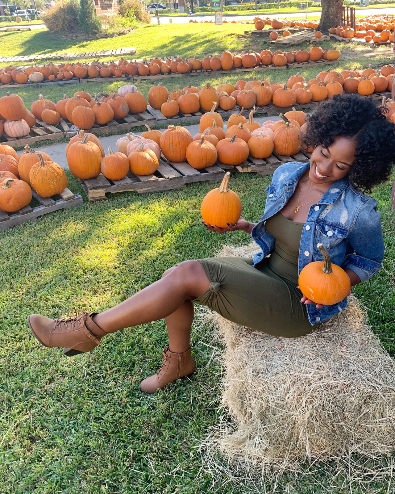 Woman in olive green dress, denim jacket and tan lace-up boots sitting on a hay bale at a pumpkin patch