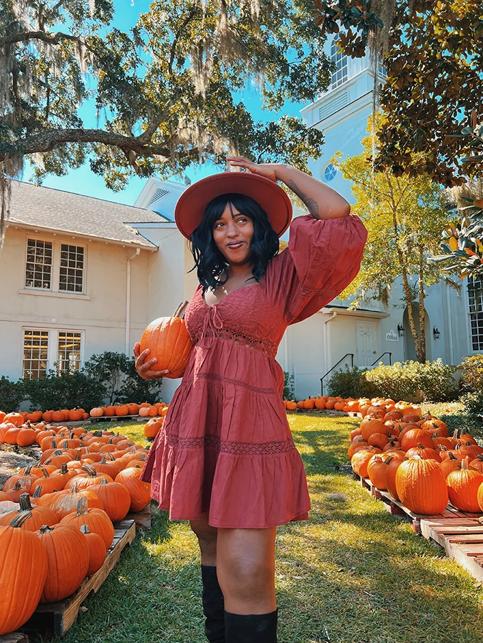 Woman in rust lace mini dress and wide brim hat with black boots at a pumpkin farm