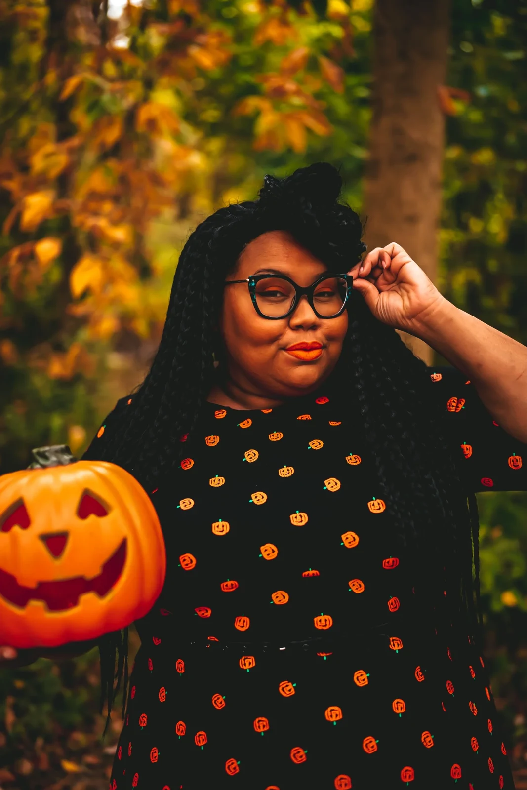 Woman in black pumpkin print dress and cat eye glasses holding a jack o lantern in autumn woods