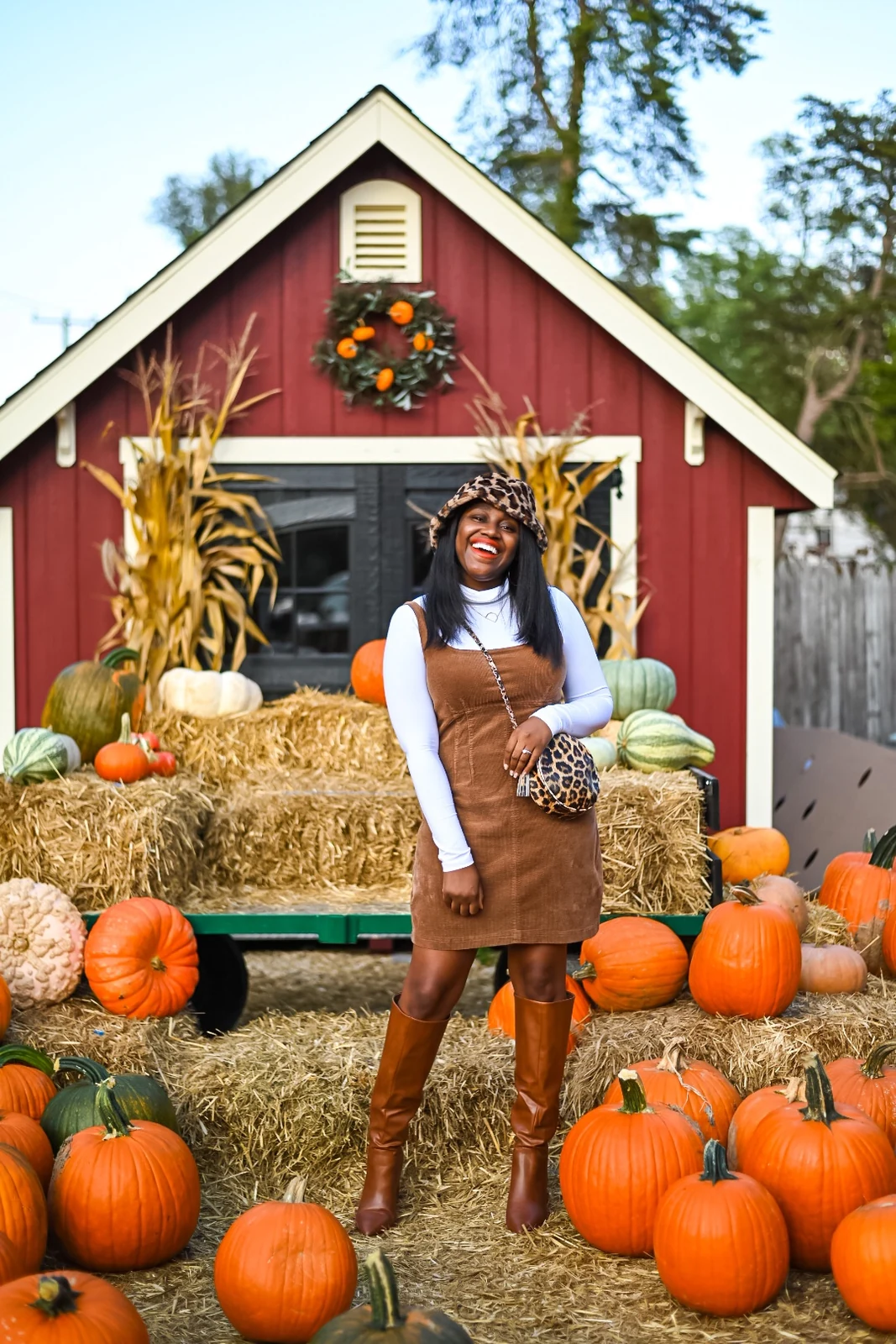 Woman in brown corduroy jumper, white turtleneck and knee high boots with leopard print hat and bag at a pumpkin patch