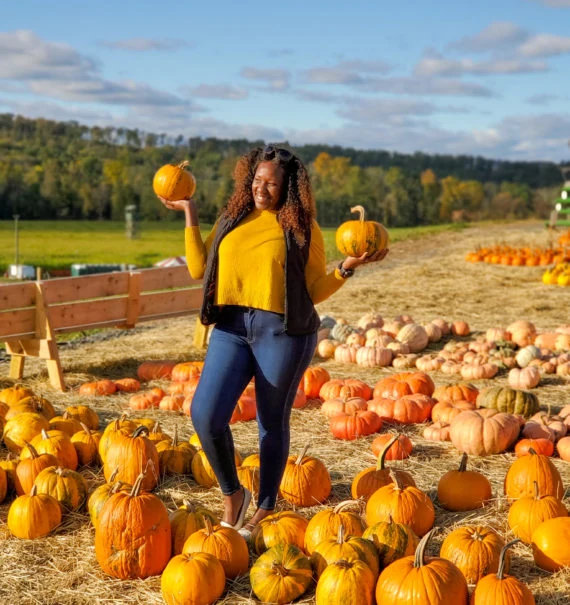 Woman in olive green long cardigan, black top and leggings with black boots holding a pumpkin