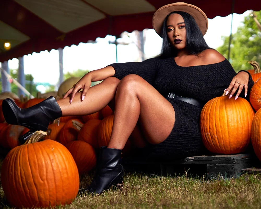Woman in black off shoulder sweater dress, tan hat and black ankle boots sitting among pumpkins