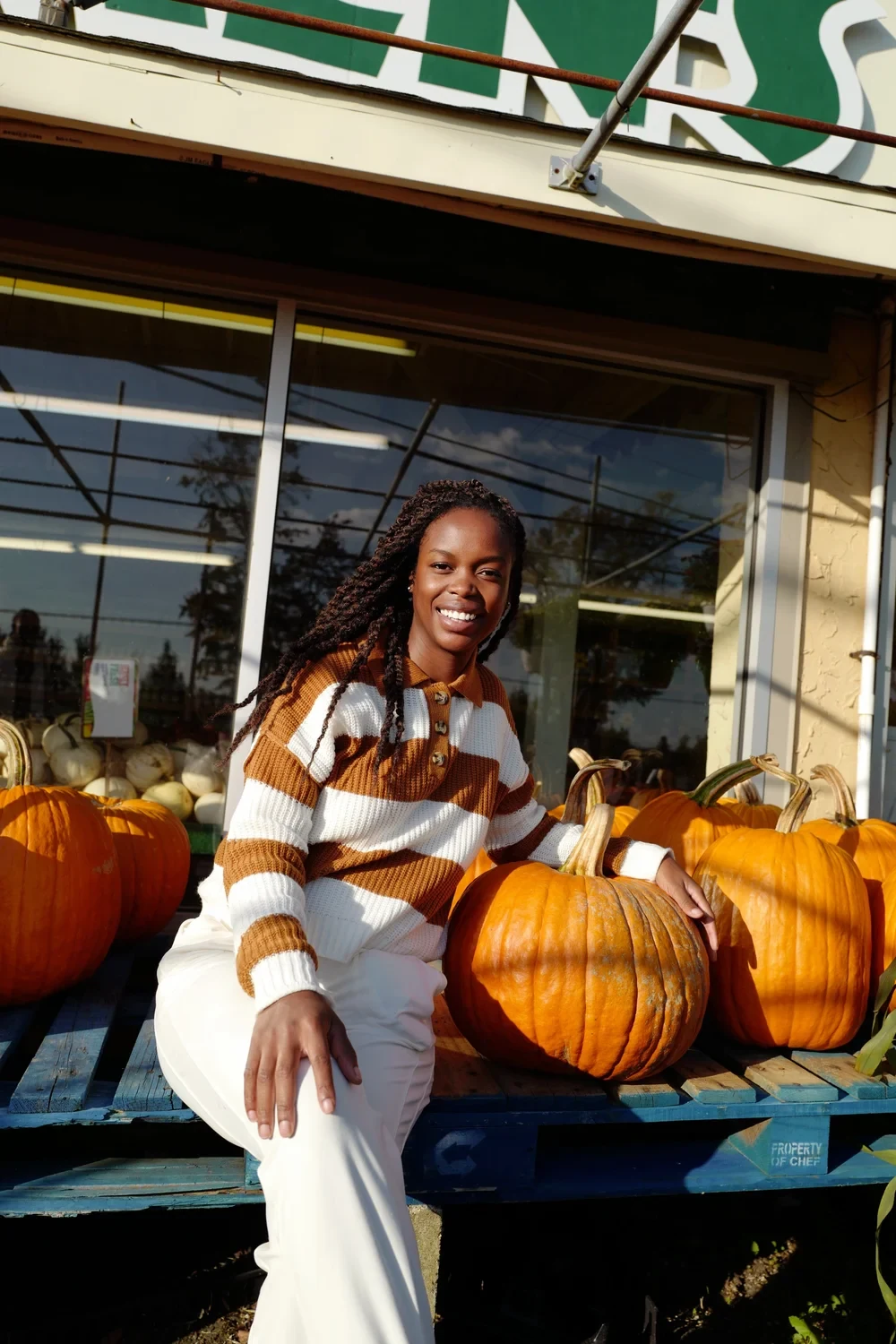 Woman in striped knit polo sweater and white pants sitting by a pumpkin stand