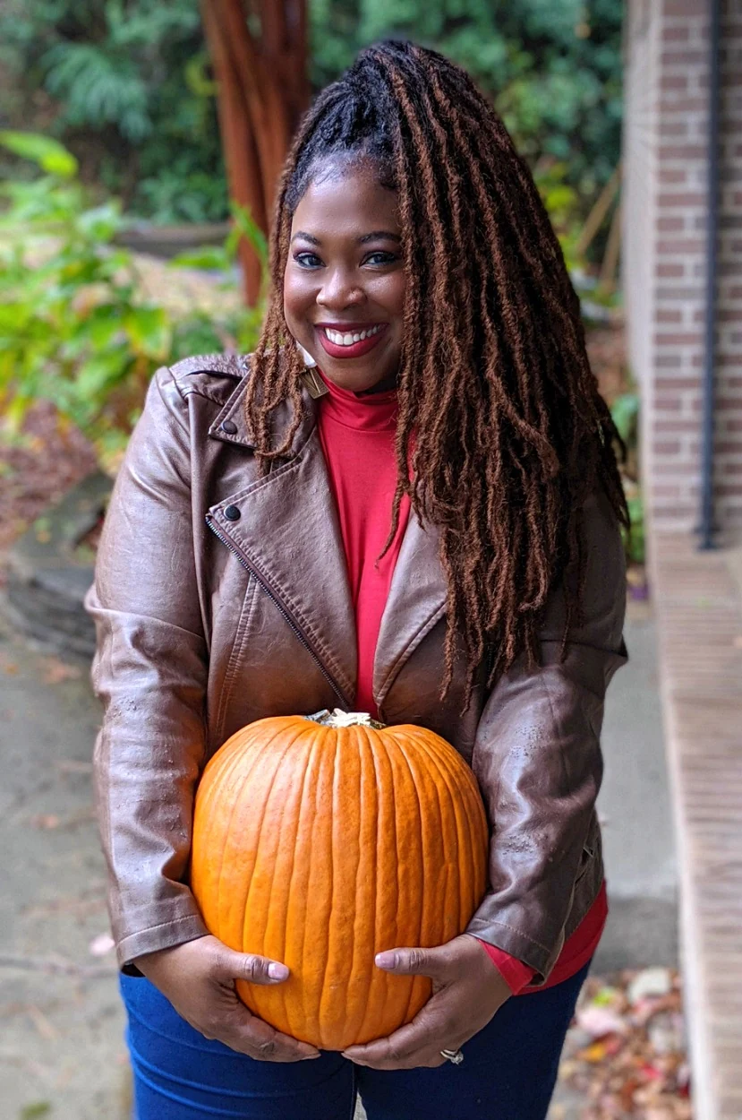 Woman in brown leather moto jacket and red top holding a pumpkin on a porch