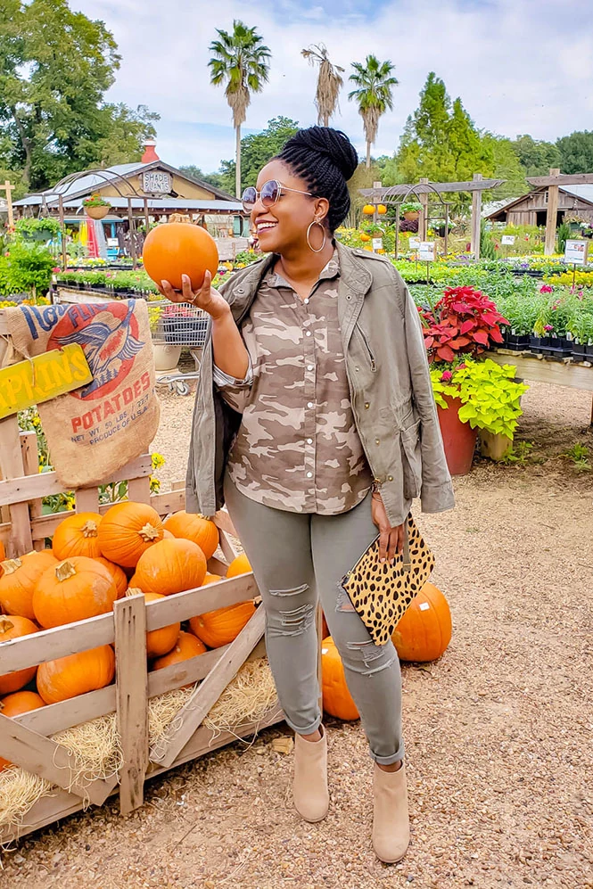 Woman in camo print shirt, olive utility jacket and ripped grey jeans with leopard clutch at a pumpkin stand