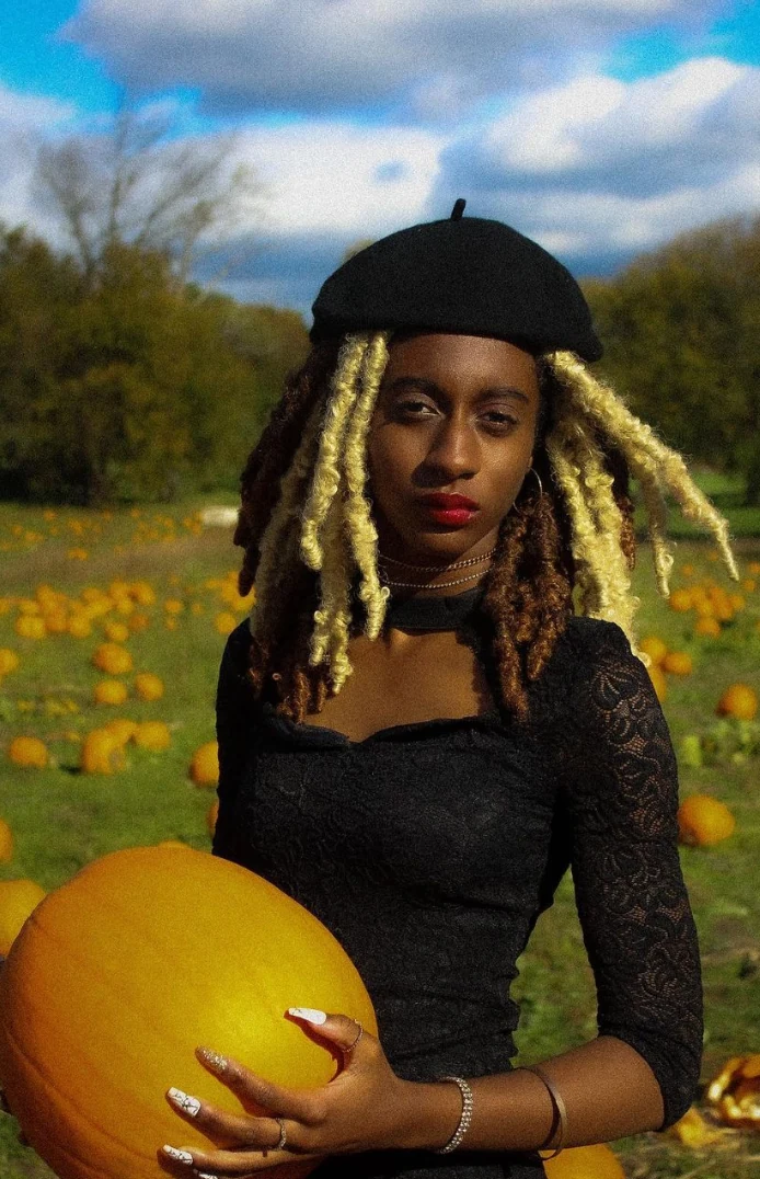 Woman in black beret and black lace top with two tone locs holding a pumpkin in a pumpkin field