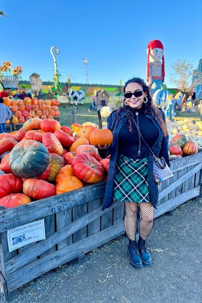 Woman in black top, green plaid skirt, fishnet tights and combat boots holding a pumpkin at an orchard