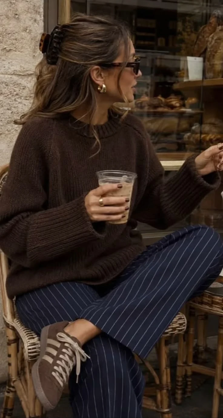 Woman in a chunky chocolate brown sweater with pinstripe trousers holding a coffee at a cafe