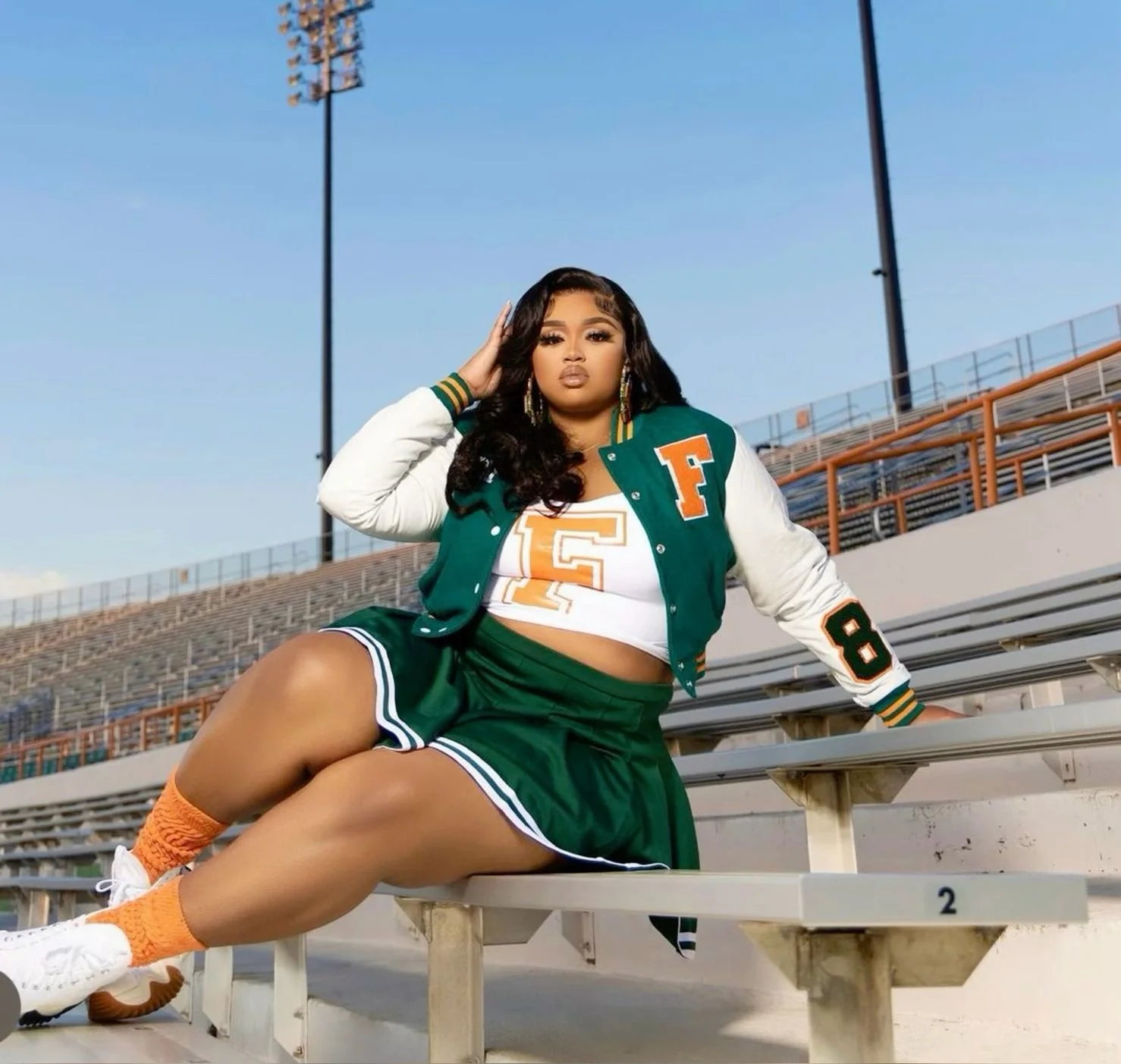 Plus size fall game day outfit: woman in a varsity letterman jacket sitting on stadium bleachers