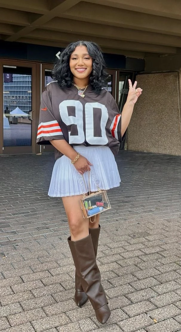 Fall game day outfit for Black women: brown number jersey with white pleated skirt and tall brown boots