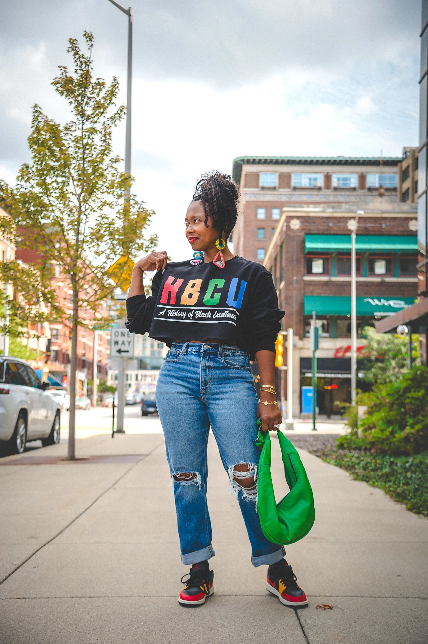 Fall game day outfit for Black women: colorful HBCU sweatshirt with ripped jeans, green bag and sneakers