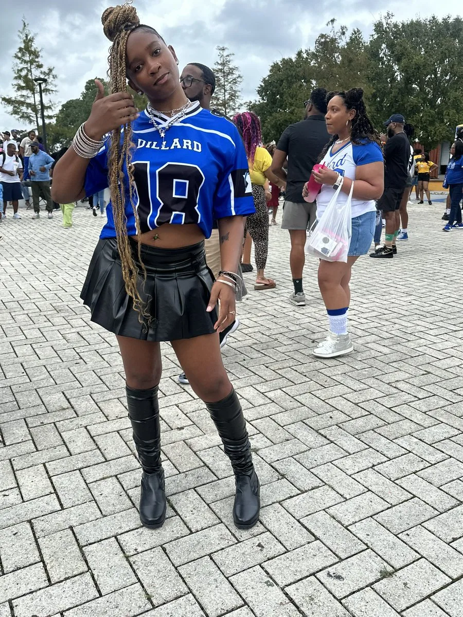 Black woman in a blue number 18 jersey and black pleated skirt with tall black boots