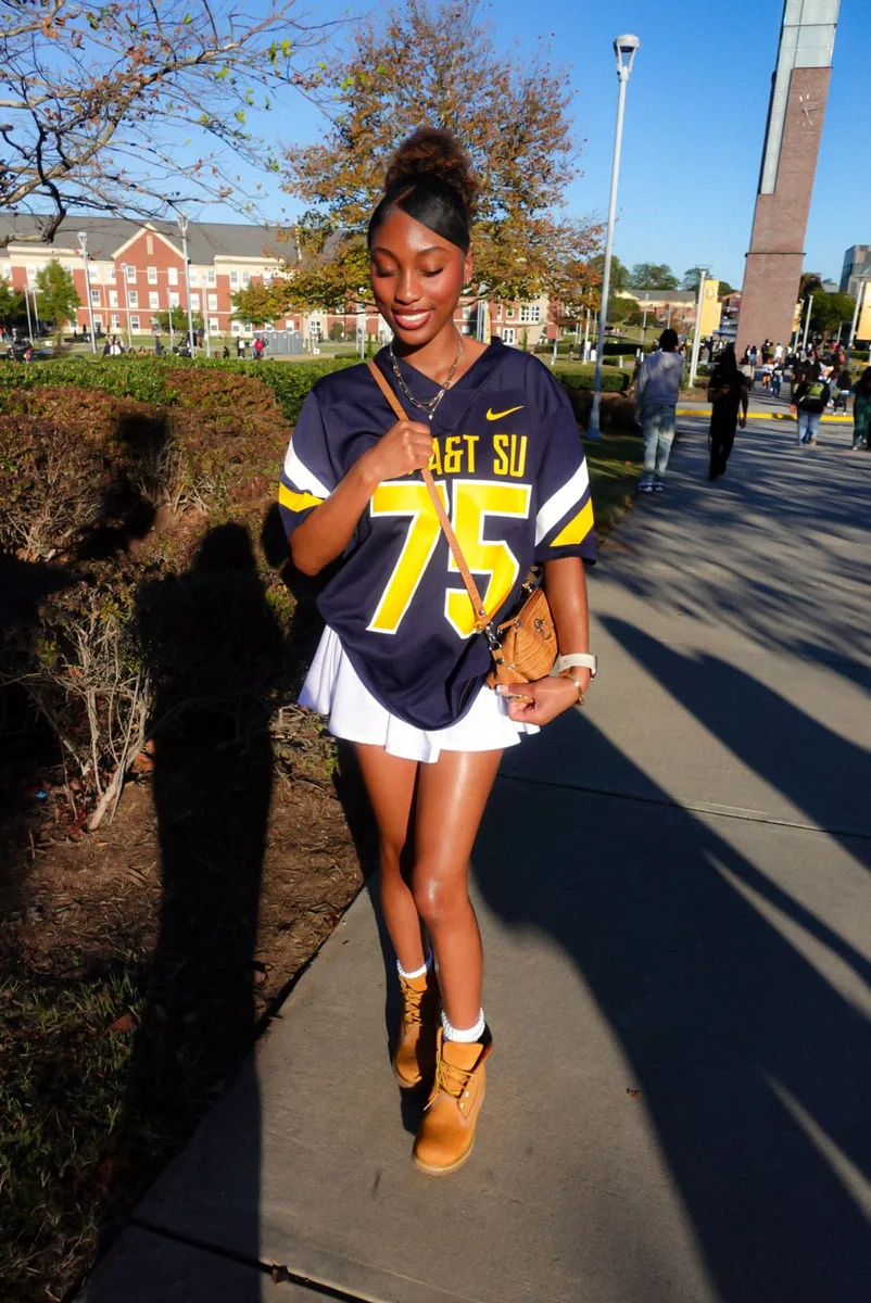 Black woman in a navy number 75 jersey and white skirt with brown boots on campus