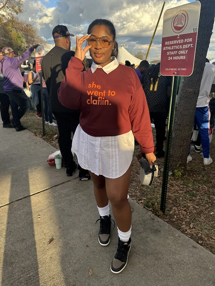 Black woman in a rust sweatshirt and white skirt with a cap at homecoming