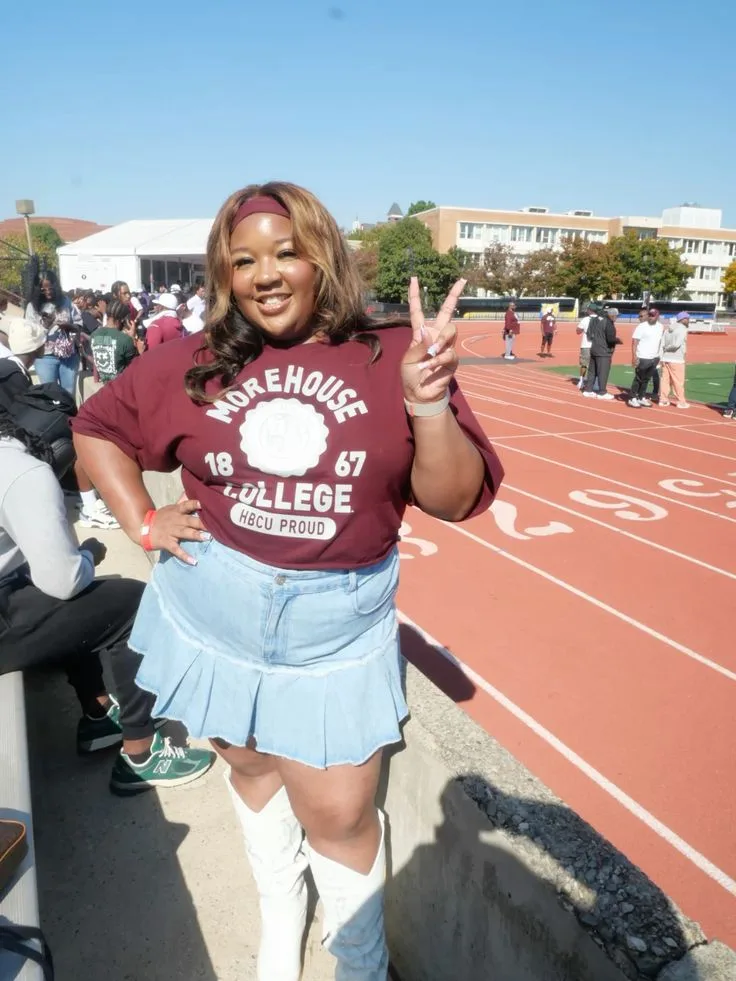 Black woman in a maroon college tee and light blue pleated skirt with white boots on a track