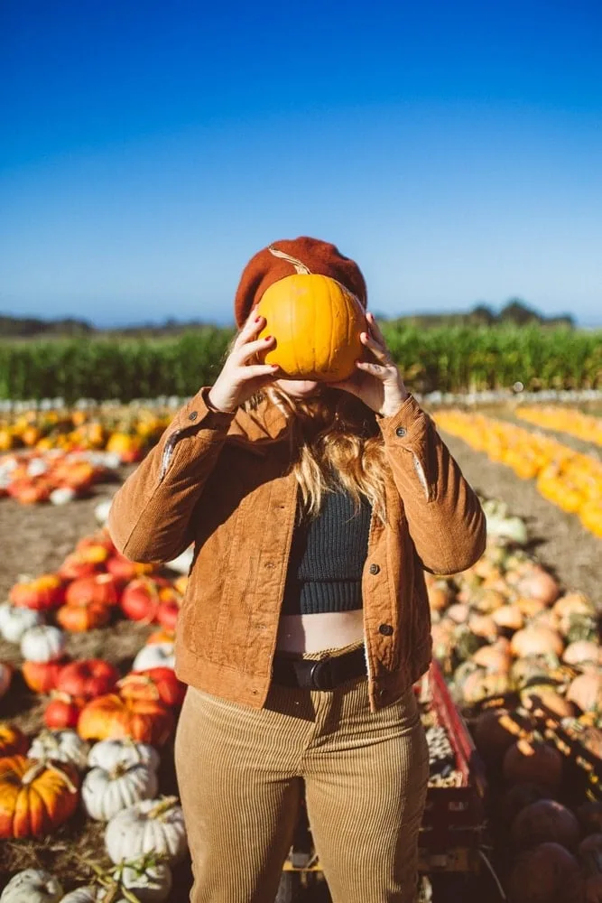Fun pumpkin patch outfit: tan suede jacket with jeans holding a pumpkin over her face