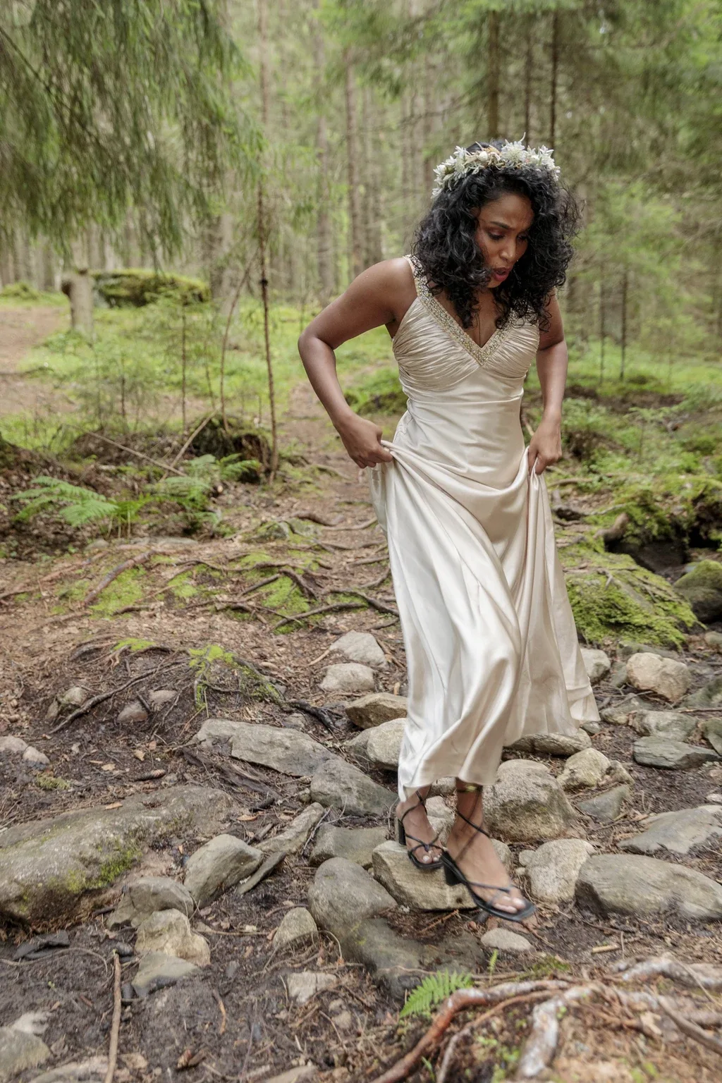 Woman in a champagne satin gown and heels crossing a rocky forest stream