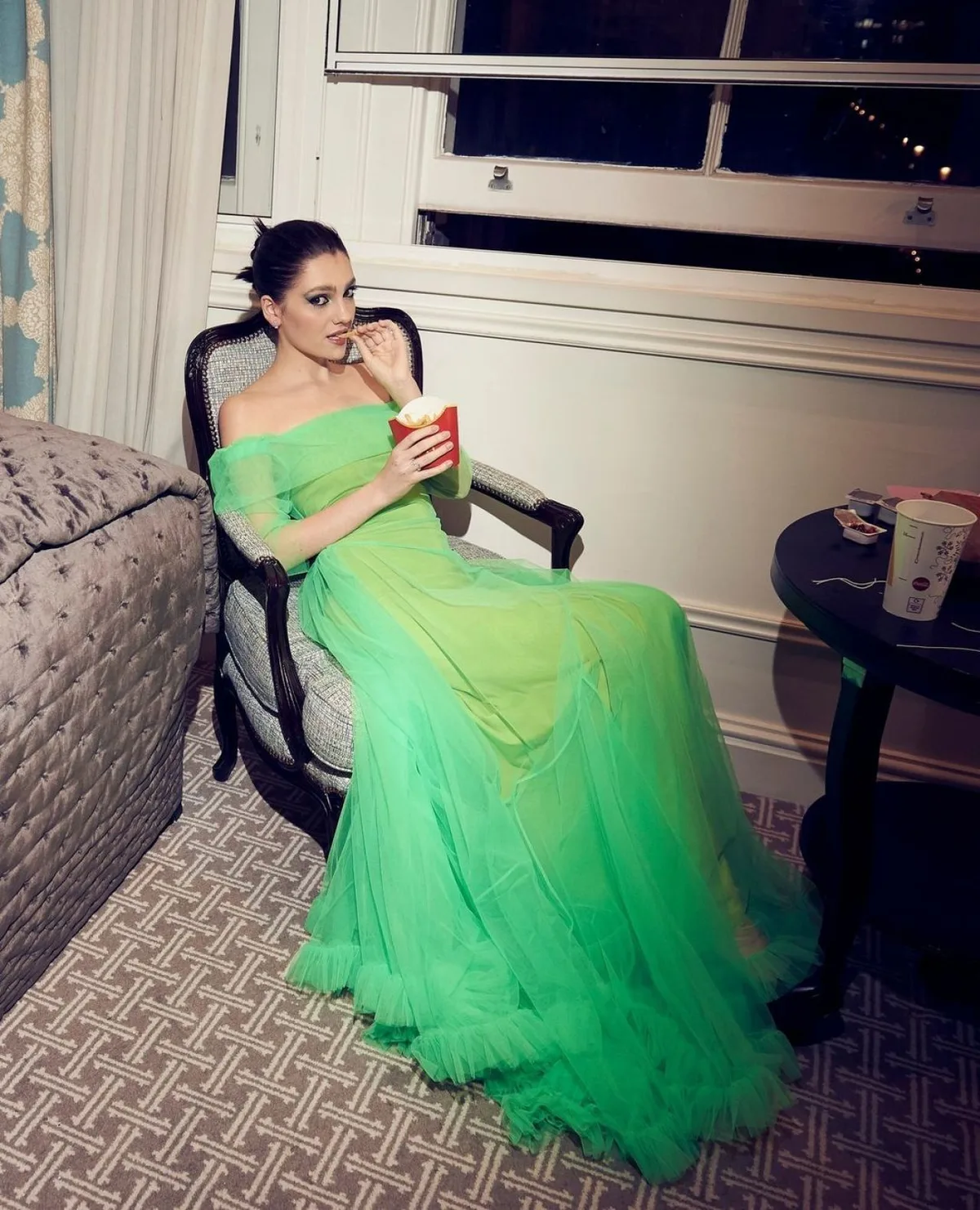 Woman in a green tulle gown eating fries in a hotel room