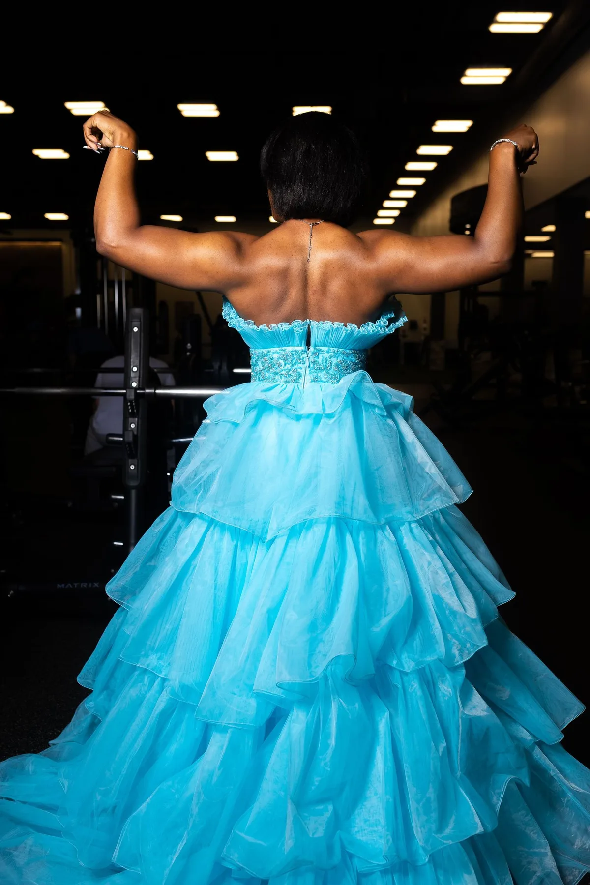 Woman in an aqua blue ballgown flexing her back muscles in a gym