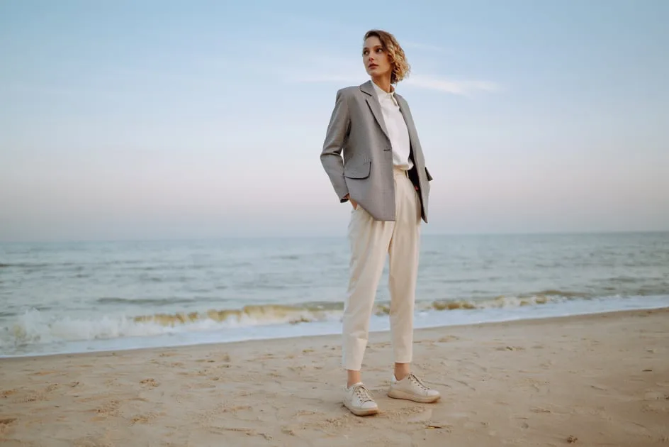Woman in a grey blazer and sneakers standing on a beach