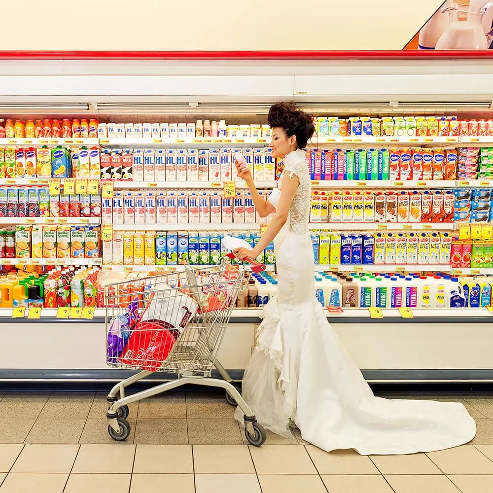 Bride in a white gown pushing a shopping cart in a grocery store dairy aisle