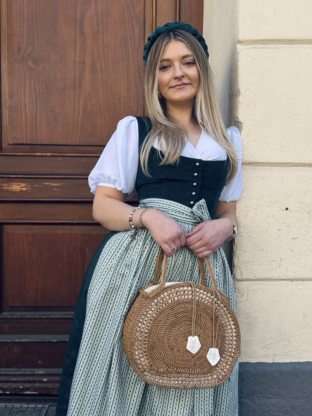 Woman in a dark green dirndl with a striped apron holding a straw bag