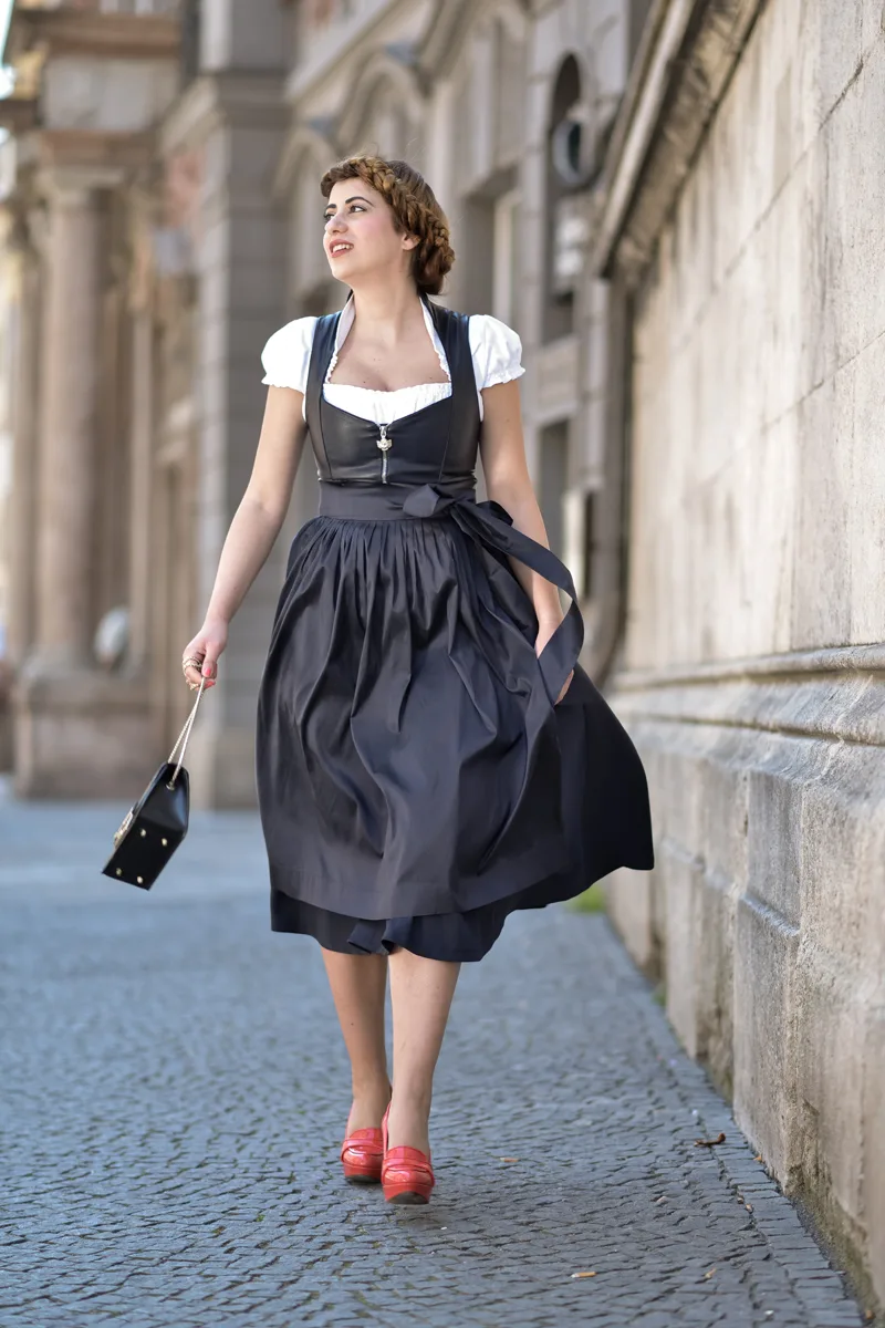 Woman in an all black dirndl with red loafers and a mini bag on a cobblestone street