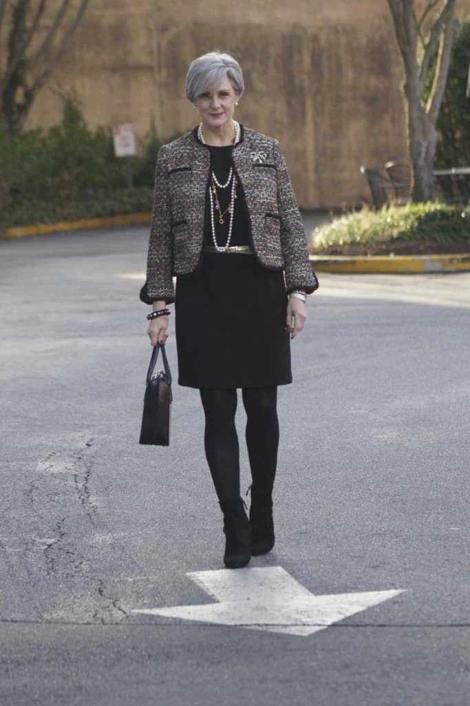 Woman over 50 wearing a tweed jacket over a black dress with tights and boots