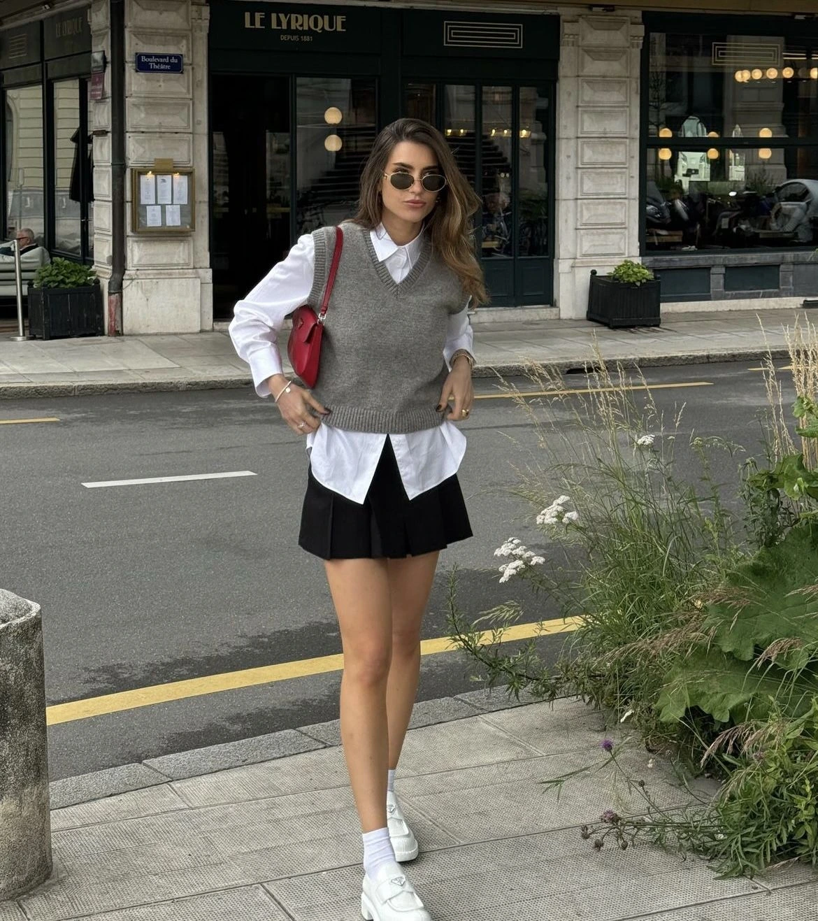Woman in a grey sweater vest over a white shirt with a black pleated mini skirt