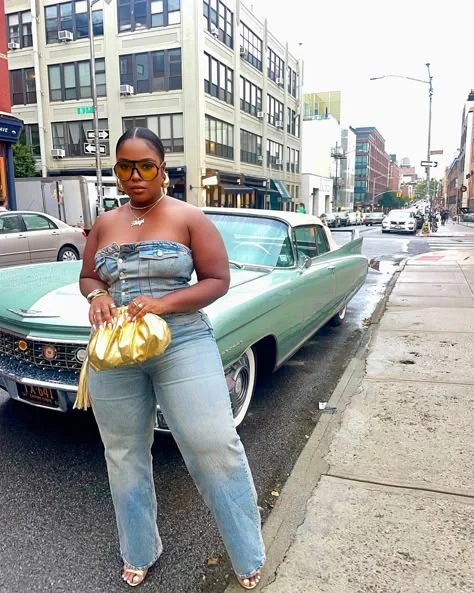 Plus size baddie in denim strapless jumpsuit holding gold clutch