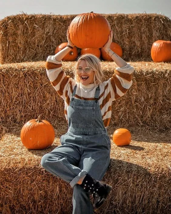 Fall pumpkin patch outfit: striped sweater under denim overalls holding a pumpkin overhead