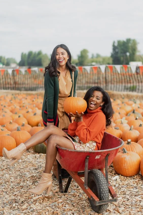 Fall pumpkin patch outfits: two women in sweaters laughing with a red wheelbarrow