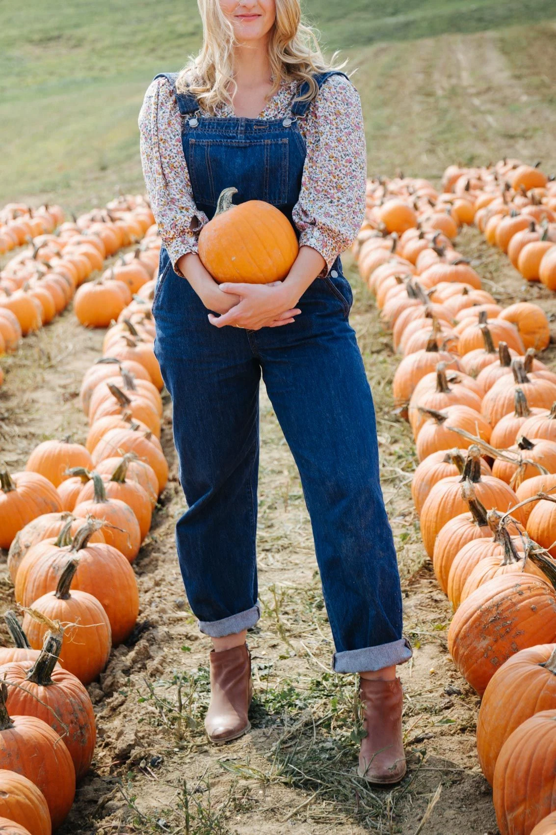 Fall pumpkin patch outfit: floral blouse layered under denim overalls holding a pumpkin