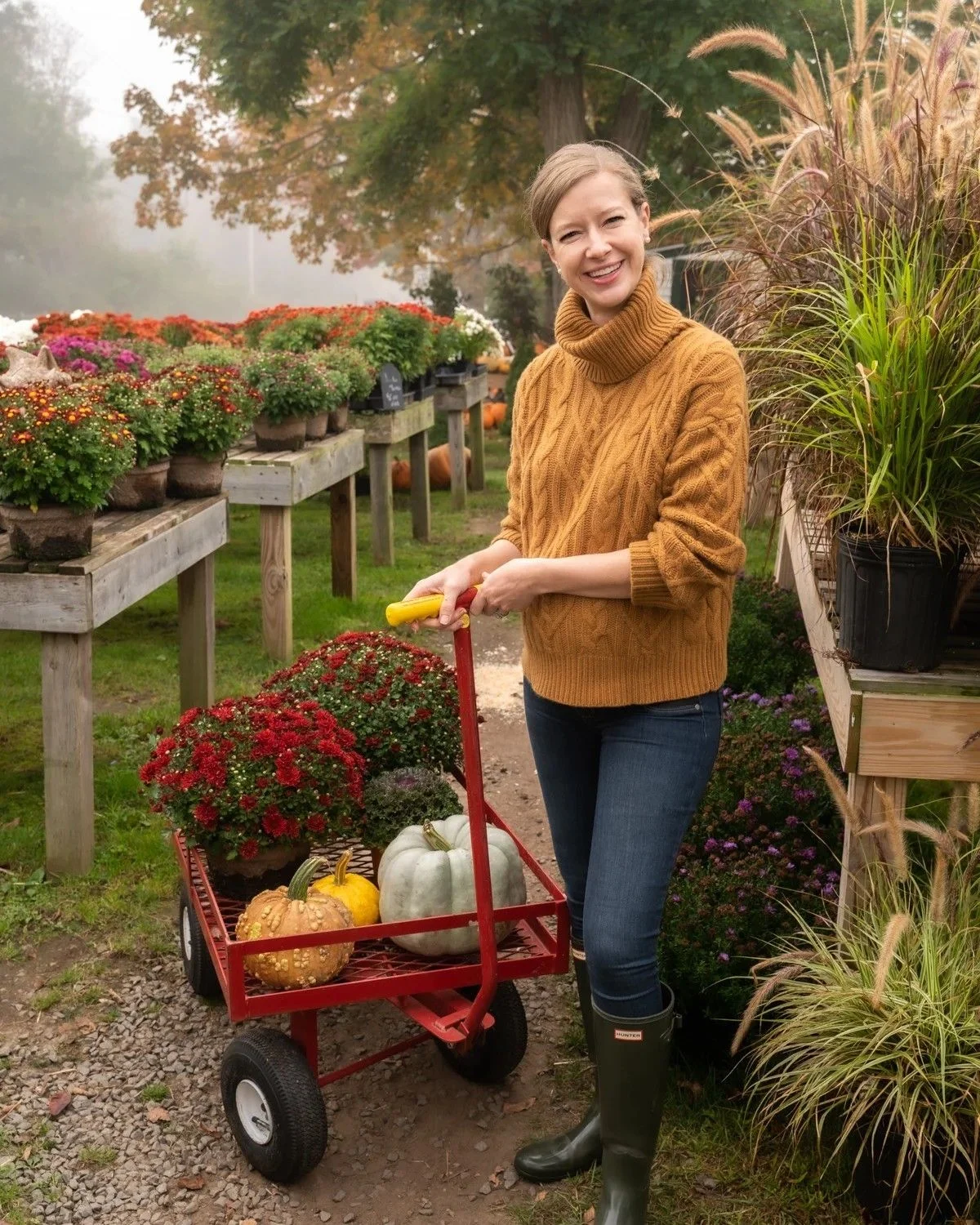 Woman in mustard cable-knit turtleneck, jeans and green wellies with a wagon of pumpkins