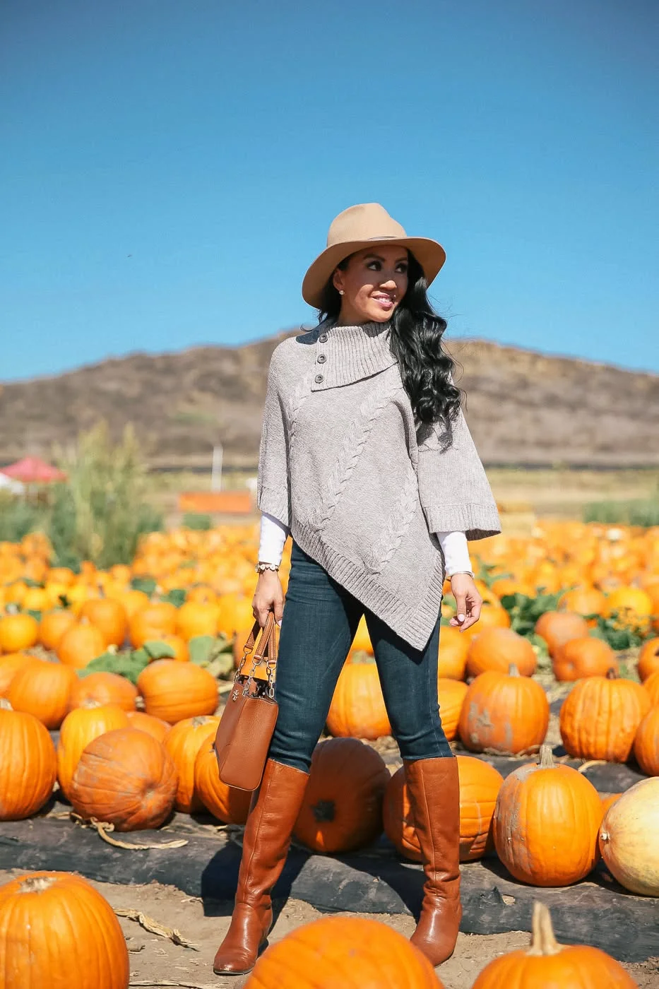 Woman in grey cable-knit poncho, dark jeans and tan boots with a felt hat in a pumpkin field