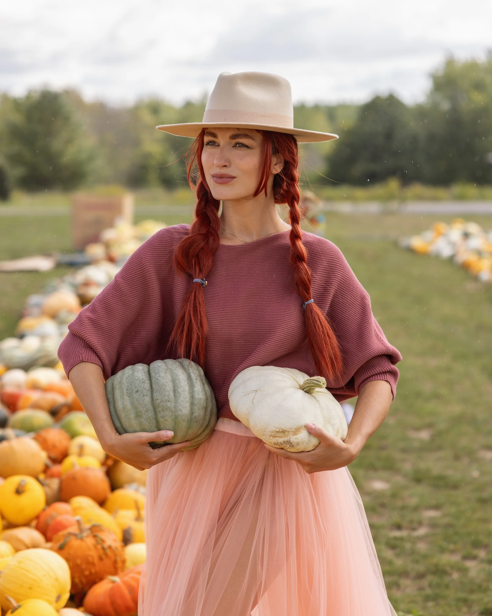 Woman in mauve sweater, blush tulle skirt and beige hat holding heirloom pumpkins