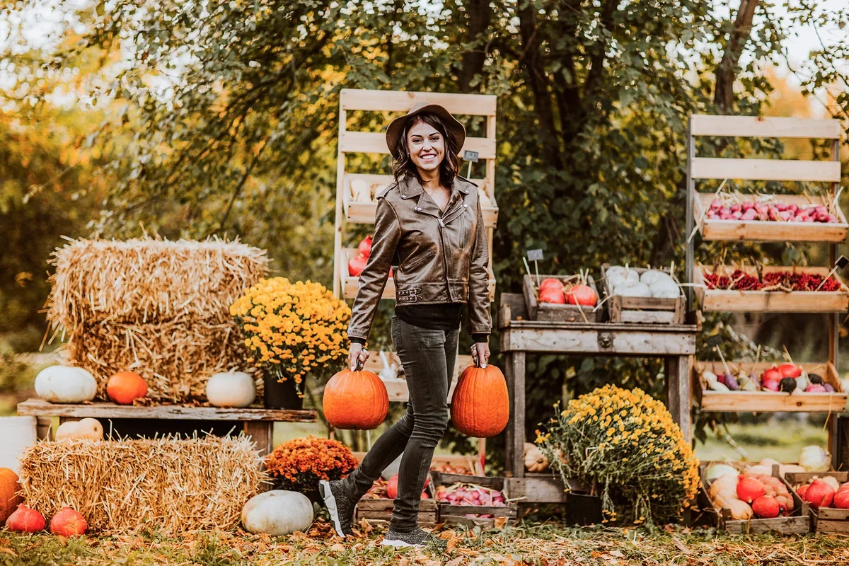 Woman in brown leather moto jacket and fedora carrying two pumpkins at a farm market