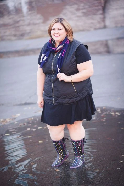 Plus-size woman in a black quilted vest, navy dress and patterned rain boots standing in a puddle
