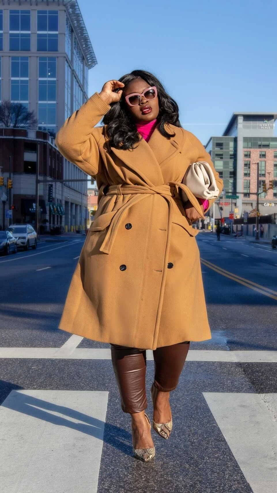 Plus-size woman in a tan trench coat, pink turtleneck and brown leather pants crossing a wet street