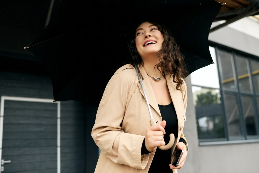 Plus-size woman in a beige trench coat holding a black umbrella on a rainy day