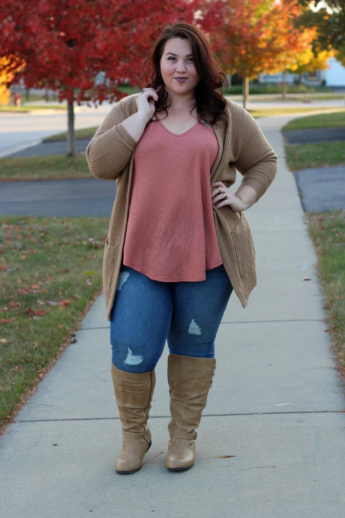 Plus-size woman in a camel coat, mauve top, ripped jeans and tan boots on an autumn sidewalk