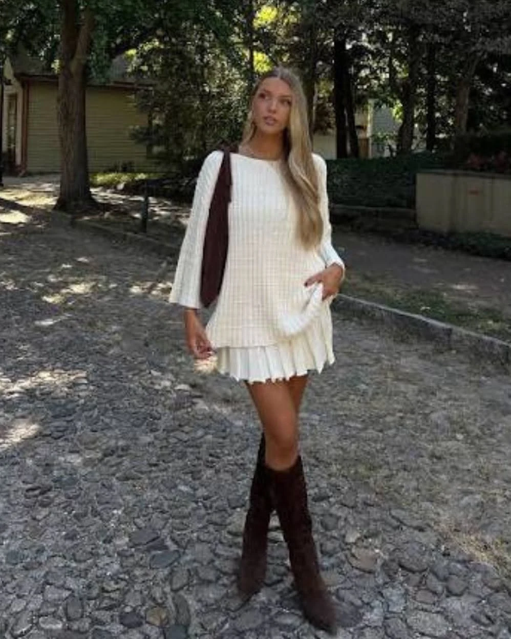 Woman in a cream knit sweater dress with brown knee high boots standing outdoors