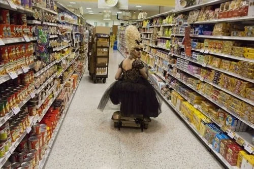 Woman in a dark gown riding a shopping cart down a store aisle